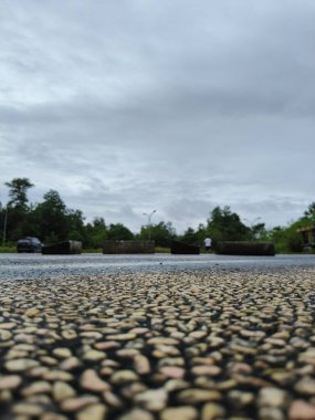 Üzerinde küçük çakıl taşları olan çakıl yolu. Yol boş görünüyor, arkada ne araba ne de insan var. Gökyüzü bulutlu ve gri. Yolun her iki tarafında da ağaçlar ve çalılar var.