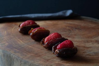 Minimalist ve karamsar bir yemek kompozisyonu. İçi taze çilekle doldurulmuş hurmalar, ahşap bir tahtanın üzerinde düzgün bir sıra halinde dizilmiş. Koyu arkaplan ve yumuşak ışıklandırma zengin bir gurme f oluşturur