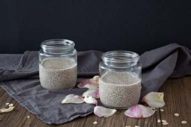 Sade chia tohumu pudingiyle dolu iki cam kavanozun temiz ve zarif bir sunumu, dağınık gül yaprakları ve yulaf lapalarıyla kaplı koyu kumaş peçetenin üzerine kurulmuş. Huysuz arka plan ve doğal dokular