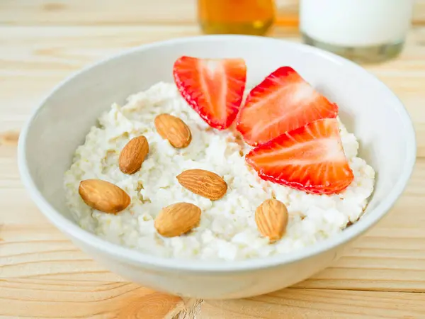 stock image A bowl of oat porridge topped with almonds and fresh strawberries served with milk and honey on wooden table. Healthy breakfast.