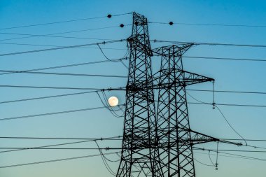 Yüksek voltajlı iletim hatlarının kuleleri gün batımında, arka planda dolunay varken.