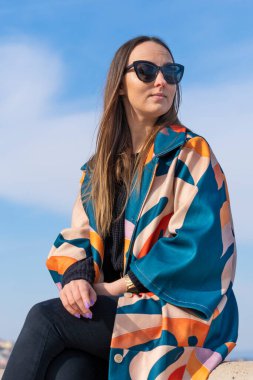 Renkli ceketli, güneş gözlüklü, arka planda mavi gökyüzü olan genç bir kadın.