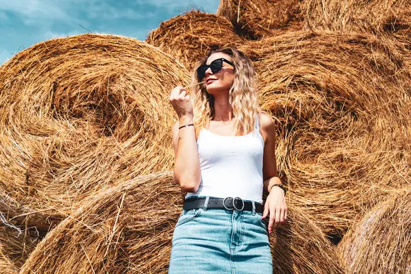 Sarışın, beyaz tenli, güneş gözlüklü, yaz güneşinin tadını çıkaran, saman yığını geçmişi olan bir kadın.