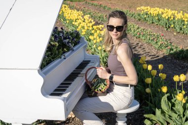 Arkasında bahar sarısı laleleri olan beyaz piyanonun başındaki sarışın kadın.