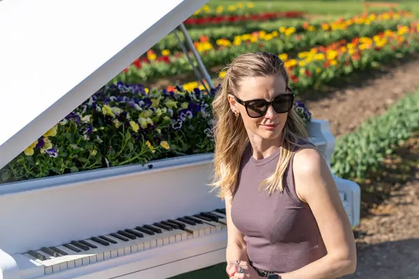 Arkasında bahar sarısı laleleri olan beyaz piyanonun başındaki sarışın kadın.