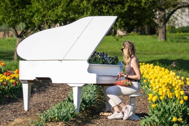 Arkasında bahar sarısı laleleri olan beyaz piyanonun başındaki sarışın kadın.