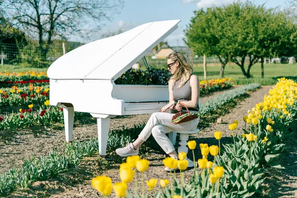 Arkasında bahar sarısı laleleri olan beyaz piyanonun başındaki sarışın kadın.