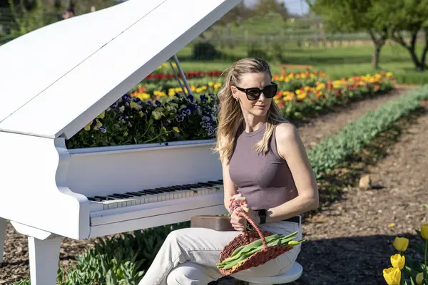 Arkasında bahar sarısı laleleri olan beyaz piyanonun başındaki sarışın kadın.