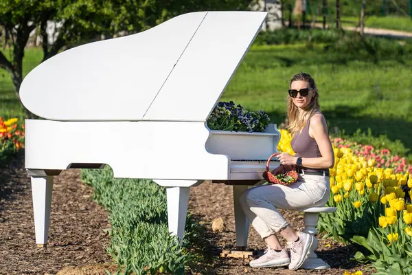 Arkasında bahar sarısı laleleri olan beyaz piyanonun başındaki sarışın kadın.
