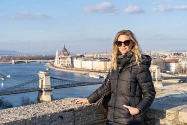 Sarışın kadın Budapeşte 'de poz veriyor. Arka planda Pest Köprüsü, Tuna Köprüsü ve parlamento binası var.