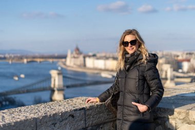 Sarışın kadın Budapeşte 'de poz veriyor. Arka planda Pest Köprüsü, Tuna Köprüsü ve parlamento binası var.