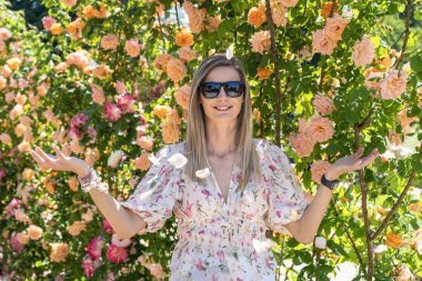 Sarışın, güneş gözlüklü bir kadın bahçede çiçek açan güllerin tadını çıkarıyor.