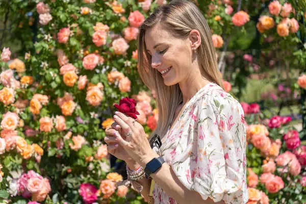 Elinde kırmızı gül olan sarışın bir kadın bahçede çiçek açan güllerin tadını çıkarıyor.