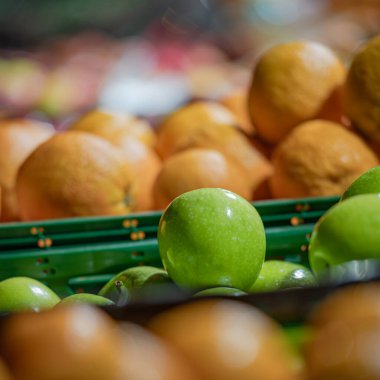 Bir markette ya da markette yeşil plastik kasalarda yeşil elma ve portakalların kare yakın çekimi. Arka planda narenciye meyveleri olan tek bir elma odaklanır..