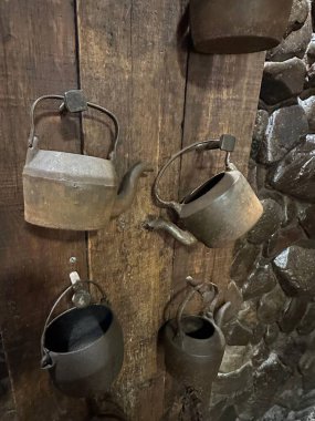 Kırsal ahşap bir duvarda asılı klasik demir çaydanlıkların sanatsal bir gösterisi. Görüntü, eski metalin dokusunu ve tahtanın doğal yapısını yakalıyor. Tarihi ve nostaljik bir atmosfer yaratıyor..