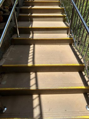 Metal tırabzanlar ve sarı güvenlik çizgileriyle aydınlık beton bir merdiven. Taş zemin boyunca ilgi çekici gölgeler bırakıyor. Geometrik simetri ve çevresel bileşimi yakalıyor..