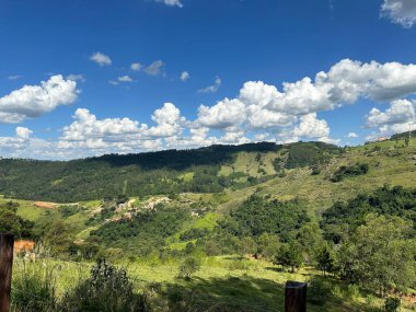 Bereketli yeşil tepelerin panoramik manzarası yoğun orman parçalarıyla kaplı canlı mavi bir gökyüzünün altında tüylü beyaz bulutlarla dolu, huzurlu ve huzurlu bir manzara yaratıyor..
