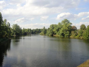 Sakin sularla çevrili sakin bir nehir manzarası bulutlu bir gökyüzü altında yemyeşil ağaçlarla çevrili, sükuneti ve doğanın güzelliğini çağrıştıran..