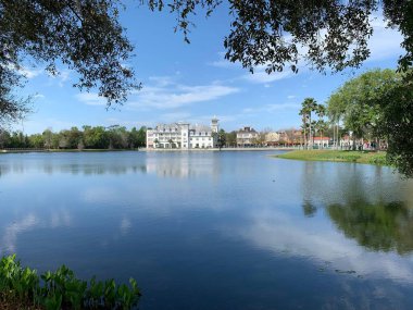 Bereketli yeşilliklerle çevrili, uzakta lüks bir tatil köyü olan huzurlu bir göl manzarası parlak mavi gökyüzünün altındaki sakin sulara yansıyor..