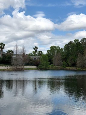 Sakin sularla gölün sakin manzarası bulutlu mavi gökyüzünü yansıtıyor. Etrafı yemyeşil ağaçlarla çevrili ve bazı çıplak ağaçlarla çevrili. Sakin bir doğa sahnesi..