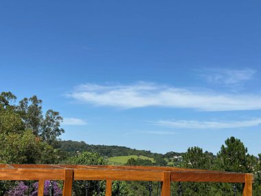Açık mavi gökyüzü ve yeşil tepeleri ve yemyeşil ağaçları olan nefes kesici bir panoramik manzara, ön plandaki ahşap korkulukla tamamlanmış, sakin bir atmosfer yaratıyor..
