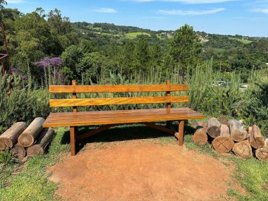 Sunlit wooden bench in a lush green park surrounded by logs and vibrant vegetation, offering a serene view of distant hills and a blue sky, evoking a sense of tranquility and natural beauty. clipart