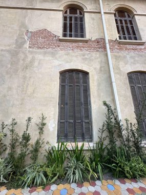 Yıpranmış duvarları, ahşap pencereleri ve çevresindeki bitki örtüsü olan tarihi bir binanın dış görünüşü. Kırsal duvarla pencerelerin simetrisi arasındaki zıtlık nostalji ve tarihi çağrıştırıyor..