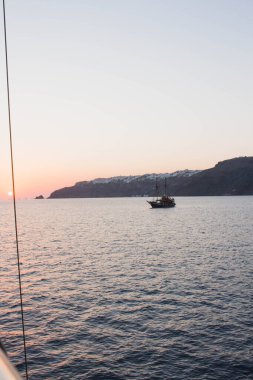 Santorini Yunanistan 'da sıcak bir yaz gününde, Ege Denizi' nde güneş ufukta batıyor ve bir tekne uzaklarda seyrediyor. Kasaba yakındaki adalarda zar zor görülebiliyor ama su sakin ve güzel..
