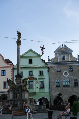 A female slackliner demonstrates her skills and daring tricks on a rope above a historic European square, framed by colorful facades and a monumental statue. clipart