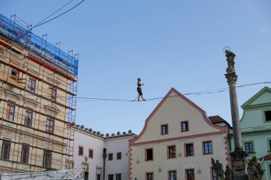 Bir kadın kaytarıcı yeteneklerini ve cüretkar hilelerini, renkli yüzler ve anıtsal bir heykel tarafından çerçevelenmiş tarihi bir Avrupa meydanının üzerinde bir iple sergiliyor..