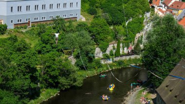 İki dağcı, tarihi binalar ve kanocularla çevrili yemyeşil bir vadide, nehrin üzerinde uzanan iplerde gösteri yapıyorlar. Manzaranın tadını çıkarıyorlar..