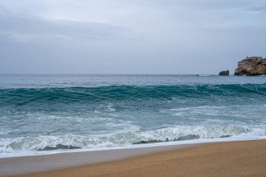 Okyanus dalgalarının yakın çekimi arka planda kayalıklar olan kumlu bir sahile nazikçe çarpıyor. Doğa, dinginlik ve sahil güzelliği teması için mükemmel bir görsel..