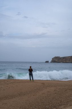 Bir kadın kumlu bir plajda duruyor, dalgalarla ve engebeli bir sahil kayalıklarıyla karşı karşıya. Seyahat, yalnızlık ve doğa temalı içerik için mükemmel..