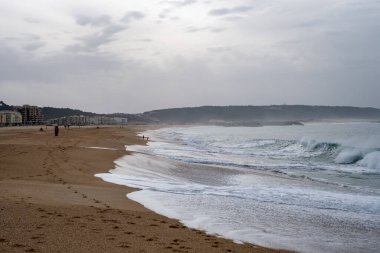 Geniş kumlu sahil, bulutlu bir gökyüzünün altında uzak kentsel binalar ve yumuşak okyanus dalgaları boyunca uzanıyor. Seyahat, turizm ve sahil manzarası temaları için mükemmel