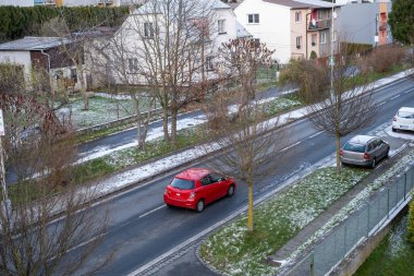 Hafif karla kaplanmış, park halindeki arabaların, çıplak ağaçların ve hareketli kırmızı bir arabanın bulunduğu bir mahalle caddesi. Kış, şehir ve banliyö temaları için mükemmel