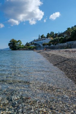 Gün ışığını yansıtan şeffaf suyla çakıl taşı plajının düşük açılı görüntüsü. Kıyı şeridi, beyaz binalar ve bulutlu mavi gökyüzünün altında yemyeşil bir kıyı köyüne doğru uzanıyor..