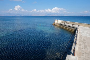 Sakin mavi denize uzanan beton iskele insanların kıyı manzarasının keyfini çıkarıyor. Temiz su, su altındaki dokuları ortaya çıkarırken uzak dağlar ve bulutlar manzaralı bir zemin oluşturur..