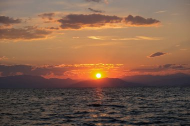 Çarpıcı bir günbatımı, göğü turuncu ve kırmızıya boyar ve dalgalı denizin üzerine altın bir parıltı saçar. Dağların uzak silueti bu büyüleyici kıyı sahnesine derinlik katıyor..