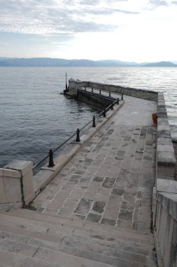 Yıpranmış bir taş iskele, suya inen bir dizi eski merdivenle sakin denize doğru uzanıyor. Gökyüzünün soluk tonları ve uzak dağlar sakin bir sahil atmosferi yaratır..