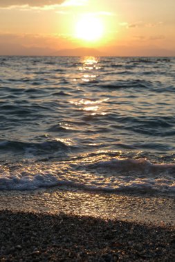 Altın güneş ufka doğru batar, dalgalı denizin üzerinde parıldayan bir parıltı bırakır. Yumuşak dalgalar çakıllı kıyıya çarpar ve büyüleyici bir ışık ve hareket etkileşimi yaratır.. 