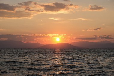 Çarpıcı bir günbatımı, göğü turuncu ve kırmızıya boyar ve dalgalı denizin üzerine altın bir parıltı saçar. Dağların uzak silueti bu büyüleyici kıyı sahnesine derinlik katıyor..