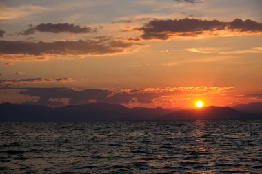 Çarpıcı bir günbatımı, göğü turuncu ve kırmızıya boyar ve dalgalı denizin üzerine altın bir parıltı saçar. Dağların uzak silueti bu büyüleyici kıyı sahnesine derinlik katıyor..