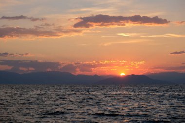 Çarpıcı bir günbatımı, göğü turuncu ve kırmızıya boyar ve dalgalı denizin üzerine altın bir parıltı saçar. Dağların uzak silueti bu büyüleyici kıyı sahnesine derinlik katıyor..