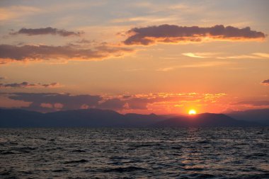 Çarpıcı bir günbatımı, göğü turuncu ve kırmızıya boyar ve dalgalı denizin üzerine altın bir parıltı saçar. Dağların uzak silueti bu büyüleyici kıyı sahnesine derinlik katıyor..