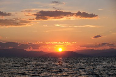 Çarpıcı bir günbatımı, göğü turuncu ve kırmızıya boyar ve dalgalı denizin üzerine altın bir parıltı saçar. Dağların uzak silueti bu büyüleyici kıyı sahnesine derinlik katıyor..