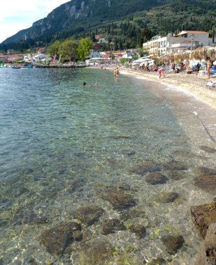 Kristal berraklığında su, güneş banyosu ve sıcak bir yaz gününün tadını çıkaran yüzücülerle dolu bir sahil. Yeşil tepeler ve büyüleyici binalar mükemmel bir sahil manzarası yaratırken, Thatched şemsiyeler kıyıda sıralanır.