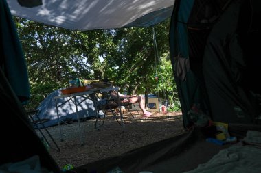 Bir çadırın içinde, ağaçların gölgesinde bir sandalyede uzanan birini gösteren huzurlu bir kamp sahnesi. Bir çadır, kamp masası ve ekipman doğada rahat bir ortam yaratır..