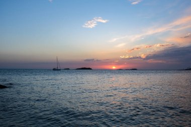 Sakin okyanusun üzerinde huzurlu bir günbatımı, küçük adaların yakınında yavaşça yüzen bir yelkenli. Gökyüzü yumuşak pastel renkleri, turuncu, pembe ve mavinin harmanlanmış tonlarını sergiler.. 