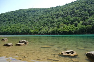 Etrafındaki yeşil orman tepelerini yansıtan berrak suyu olan sakin bir göl. Sığ sulardan büyük kayalar çıkar ve sakin doğal manzaraya derinlik katar..