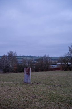 Paslı metal bir çarşaf, bulutlu bir gökyüzünün altında çimenli bir tarlada tek başına duruyor. Arka planda yapraksız ağaçlar ve yuvarlanan tepeler endüstriyel elementlerle sakin bir kırsal alan oluşturuyor..